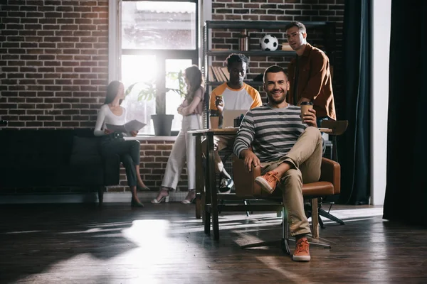 Allegro uomo d'affari seduto in poltrona con caffè per andare e sorridere alla macchina fotografica vicino a giovani colleghi multiculturali — Foto stock