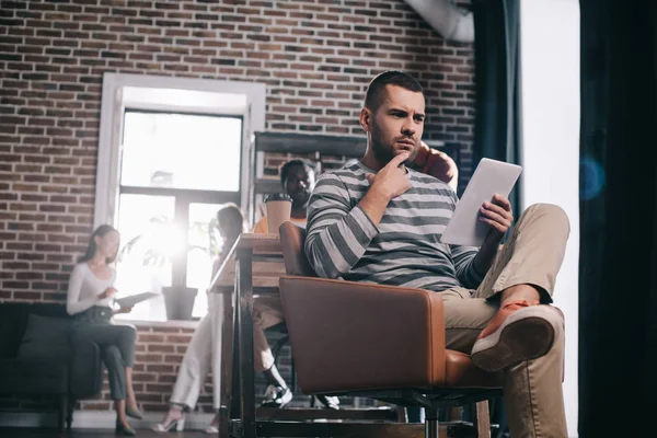 Pensativo hombre de negocios sentado en sillón y el uso de tableta digital cerca de jóvenes colegas multiculturales - foto de stock