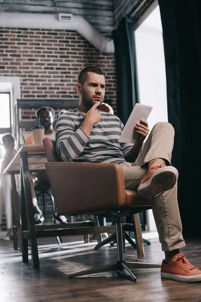 Pensativo hombre de negocios usando tableta digital mientras está sentado en sillón cerca de un colega afroamericano - foto de stock