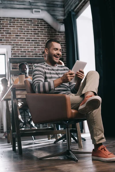 Homme d'affaires joyeux utilisant tablette numérique tout en étant assis dans un fauteuil près d'un collègue afro-américain — Photo de stock