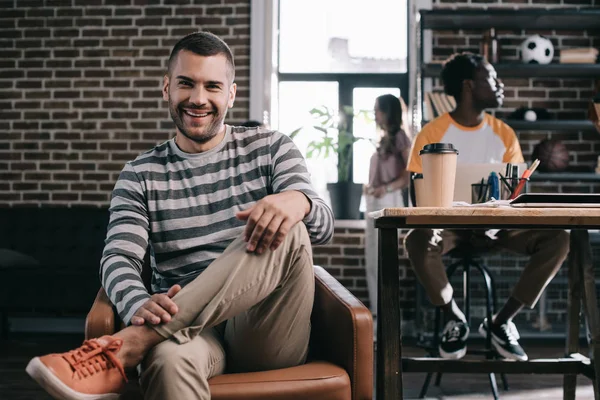 Cheerful businessman smiling at camera while sitting in armchair near young multicultural colleagues — Stock Photo