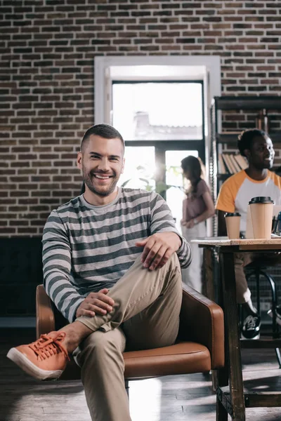 Schöner Geschäftsmann lächelt in die Kamera, während er im Sessel neben jungen multikulturellen Kollegen sitzt — Stockfoto