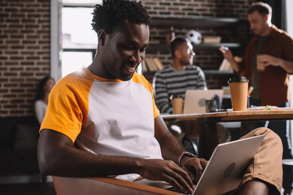 Улыбающийся африканский американский бизнесмен с помощью ноутбука, сидя в кресле рядом с молодыми мультикультурными коллегами — стоковое фото