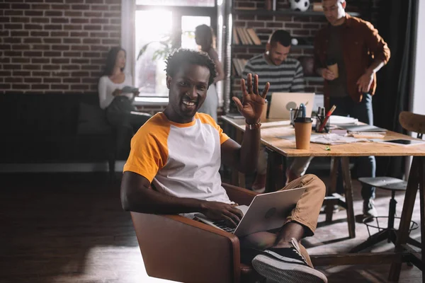 Улыбающийся африканский бизнесмен, пользующийся ноутбуком, сидя в кресле и размахивая рукой рядом с молодыми мультикультурными коллегами — стоковое фото