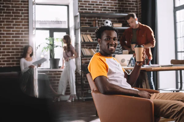Красивый африканский бизнесмен держит кофе, чтобы пойти, сидя в кресле рядом с молодыми мультикультурными коллегами — стоковое фото