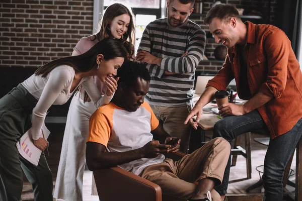 Pensativo hombre de negocios afroamericano usando teléfono inteligente mientras está sentado en un sillón rodeado de colegas multiculturales sonrientes - foto de stock