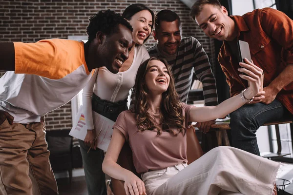 Happy businesswoman taking selfie on smartphone while sitting in armchair surrounded by cheerful multicultural colleagues — Stock Photo