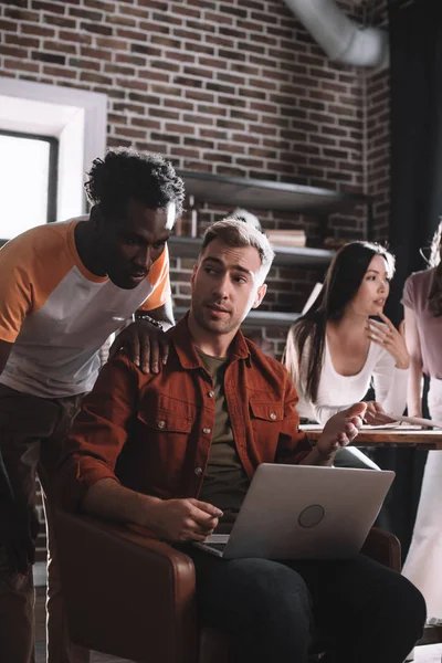 Joven hombre de negocios afroamericano de pie cerca de colega serio sentado en sillón y el uso de la computadora portátil — Stock Photo