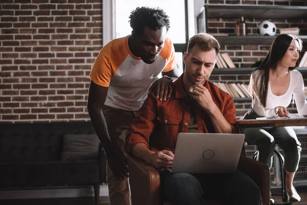 Joven hombre de negocios afroamericano de pie cerca de colega reflexivo sentado en sillón y el uso de la computadora portátil - foto de stock