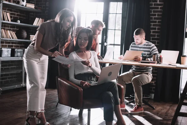Молодая предпринимательница показывает документ коллеге, сидящему в кресле с ноутбуком рядом с коллегами — стоковое фото