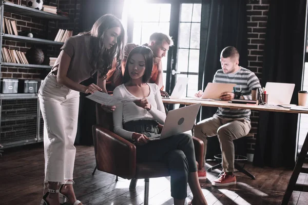 Joven empresaria mostrando documento a colega sentado en sillón con portátil cerca de colegas - foto de stock