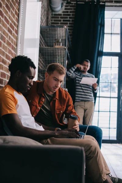 Nachdenklicher afrikanisch-amerikanischer Geschäftsmann nutzt Laptop, während er auf Sofa neben Kollege sitzt — Stockfoto