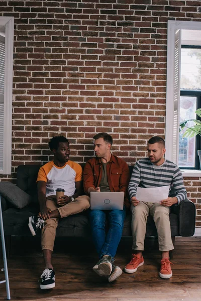 Jóvenes hombres de negocios multiculturales sentados en el sofá cerca de la pared de ladrillo y discutir ideas de negocios - foto de stock