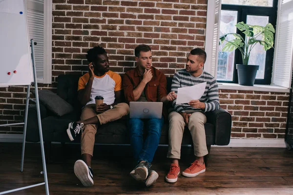 Jeunes hommes d'affaires multiculturels assis sur un canapé près d'un mur de briques et discutant d'idées d'affaires — Photo de stock