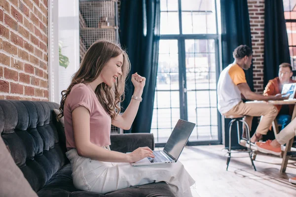 Young, happy businesswoman showing winner gesture while sitting on sofa and using laptop near multicultural colleagues — Stock Photo