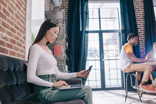 Mujer de negocios reflexivo uso de la computadora portátil mientras está sentado en el sofá cerca de colegas multiculturales — Stock Photo