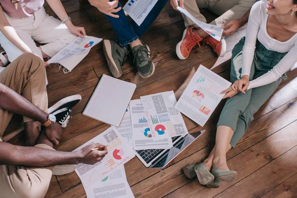 Vue recadrée de jeunes gens d'affaires assis sur le sol près de documents avec infographies — Photo de stock