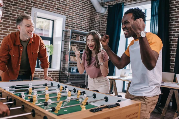 Animados empresários multiculturais mostrando gesto vencedor enquanto joga futebol de mesa no escritório — Fotografia de Stock