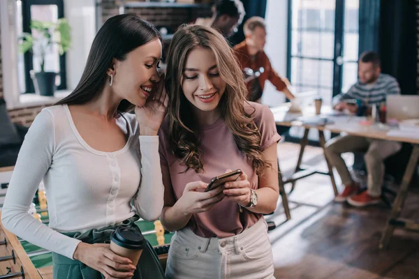 Привлекательная, улыбающаяся деловая женщина с помощью смартфона рядом с веселым коллегой держит кофе, чтобы пойти — стоковое фото