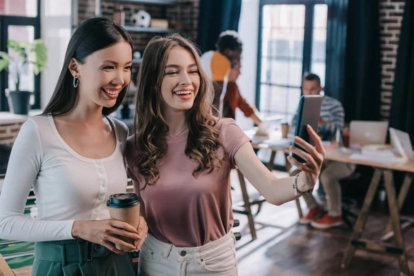 Young, cheerful businesswoman taking selfie on smartphone with cheerful colleague holding coffee to go — Stock Photo