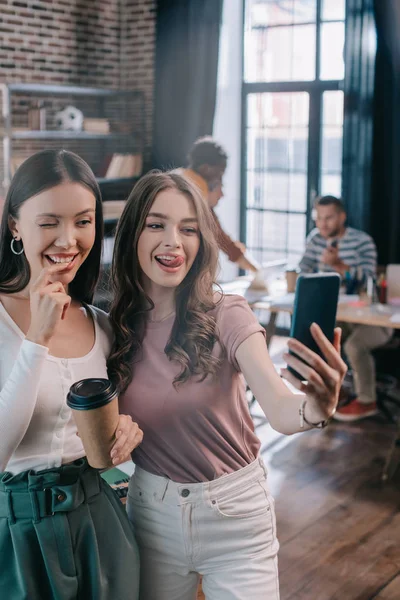 Cheerful businesswoman sticking out tongue while taking selfie on smartphone with cheerful colleague winking and holding coffee to go — Stock Photo