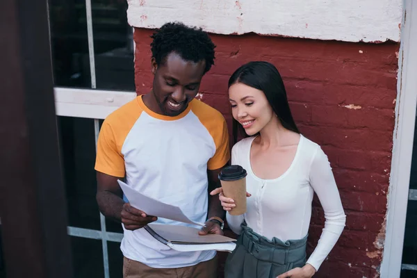 Alegre afroamericano empresario mostrando documentos a atractivo colega sosteniendo café para ir - foto de stock