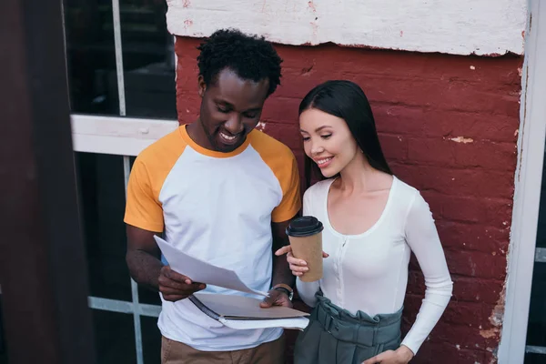 Fröhlich afrikanisch-amerikanischer Geschäftsmann zeigt attraktiven Kollegen Dokumente mit Coffee to go — Stockfoto