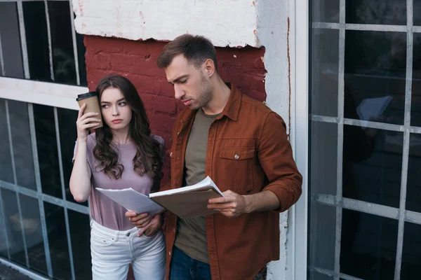 Tired businesswoman holding coffee to go near head while standing near thoughtful colleague with documents — Stock Photo