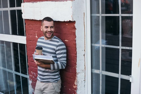 Young cheerful businessman looking away while holding documents and coffee to go — Stock Photo