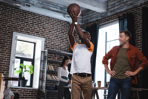 Empresário americano africano alegre segurando basquete acima da cabeça enquanto conversa com colega de pé com as mãos no quadril — Fotografia de Stock