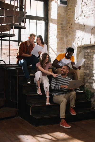 Jóvenes y sonrientes empresarios multiculturales hablando mientras están sentados en las escaleras - foto de stock