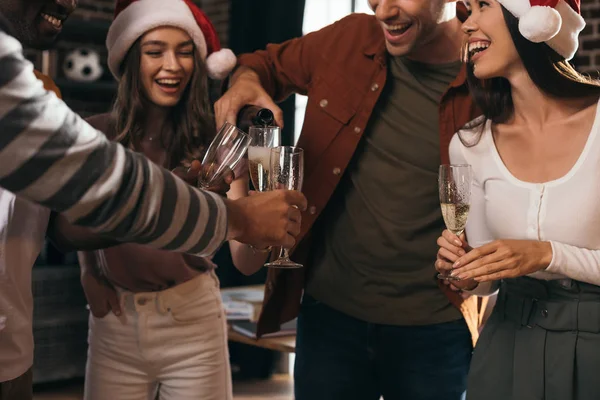 Cropped view of businessman pouring champagne in glasses of happy colleagues in santa hats — Stock Photo