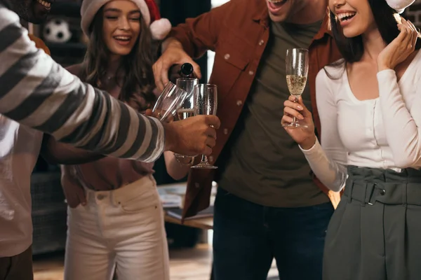 Partial view of businessman pouring champagne in glasses of happy colleagues in santa hats — Stock Photo