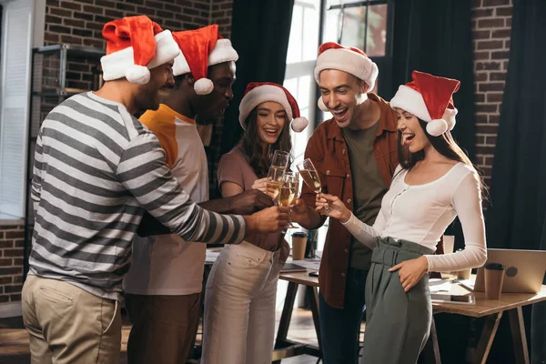 Felici uomini d'affari multiculturali in cappelli di Babbo Natale clinking bicchieri di champagne in ufficio — Foto stock