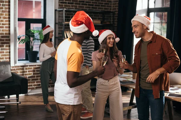 Felices empresarios multiculturales en sombreros de Santa Claus hablando mientras sostienen copas de champán en la oficina - foto de stock