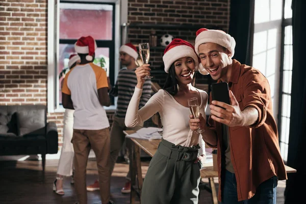 Cheerful businessman and businesswoman in santa hats taking selfie on smartphone while holding champagne glasses — Stock Photo