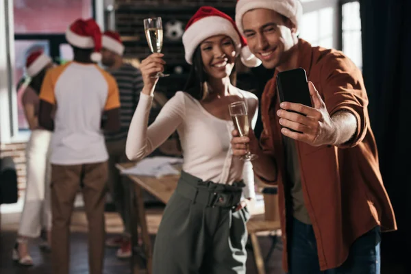 Selective focus of cheerful businessman and businesswoman in santa hats taking selfie on smartphone while holding champagne glasses — Stock Photo