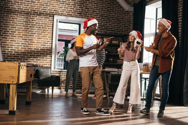 Cheerful multicultural businesspeople in santa hats talking while holding champagne glasses in office — Stock Photo