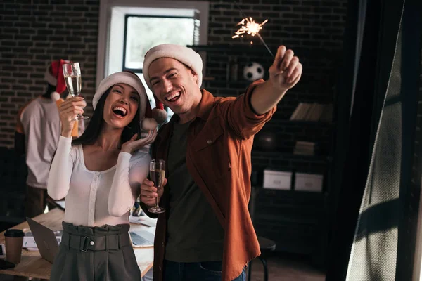 Cheerful businesspeople in santa hats looking at sparkler while holding champagne glasses — Stock Photo