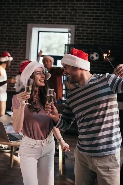 Joven, feliz hombre de negocios y mujer de negocios en sombreros de santa mirándose mientras sostiene copas de champán - foto de stock