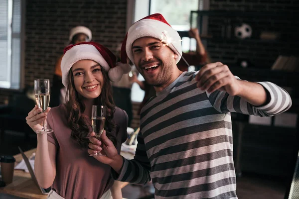 Homme d'affaires heureux et femme d'affaires dans chapeaux Santa regardant la caméra tout en tenant des verres de champagne — Photo de stock