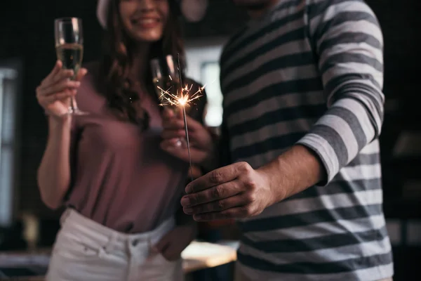 Cropped view of businessman and businesswoman in santa hats looking at sparkler while holding champagne glasses — Stock Photo