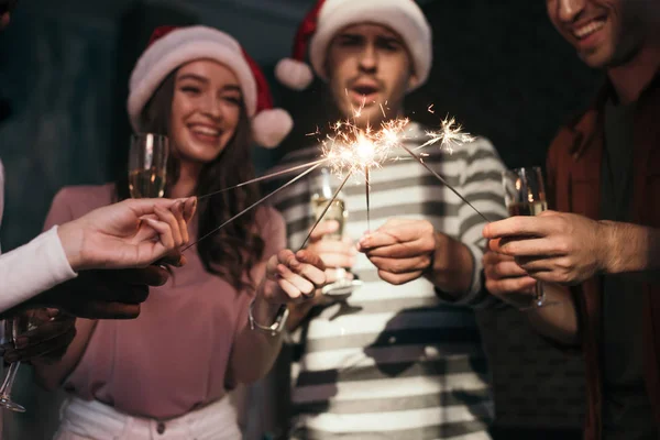 Fröhliche, junge Geschäftsleute in Weihnachtsmannhüten zünden Wunderkerzen an, während sie Champagnergläser in der Hand halten — Stockfoto