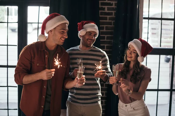 Jeunes gens d'affaires joyeux dans des chapeaux de Père Noël tenant des étincelles et des verres à champagne — Photo de stock