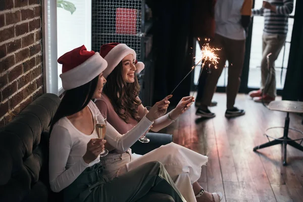 Young, happy businesswoman in santa hats sitting on sofa with sparklers and champagne glasses — Stock Photo