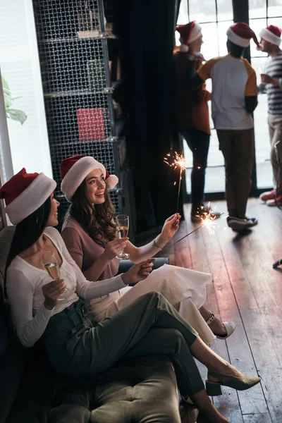 Attractive, cheerful businesswoman in santa hats sitting on sofa with sparklers and champagne glasses — Stock Photo