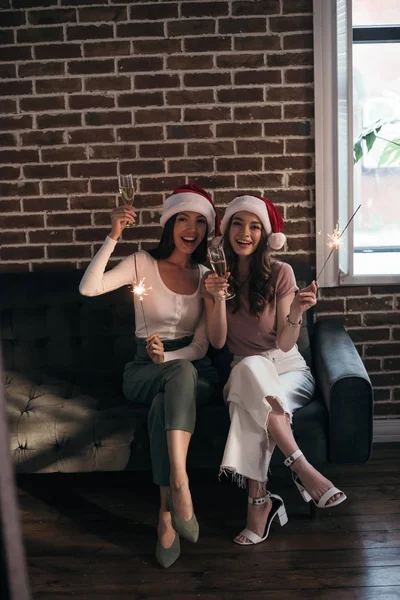 Zwei junge, glückliche Geschäftsfrauen mit Weihnachtsmannhüten, die in die Kamera lächeln, während sie mit Wunderkerzen und Sektgläsern auf dem Sofa sitzen — Stockfoto