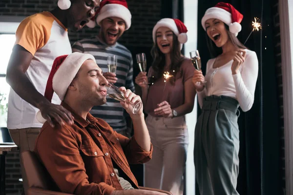 Junger Geschäftsmann trinkt Champagner, während er neben fröhlichen multikulturellen Kollegen mit Weihnachtsmannhüten steht — Stockfoto