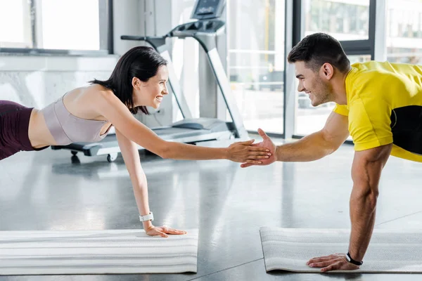 Deportista sonriente y deportista haciendo tablón y aplaudiendo en alfombras de fitness en el centro deportivo - foto de stock