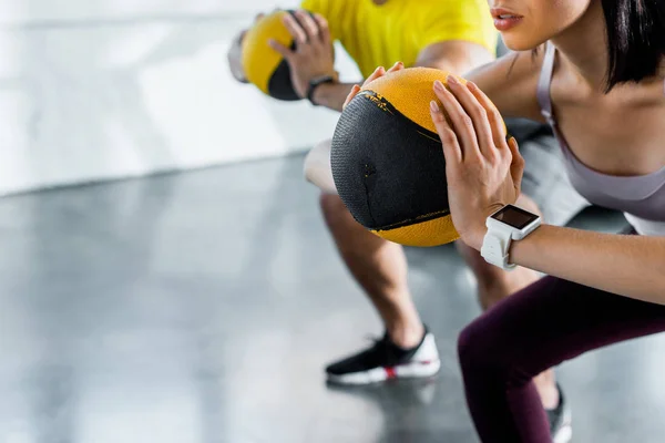 Cropped view of sportsman and sportswoman doing squat with balls in sports center — Stock Photo